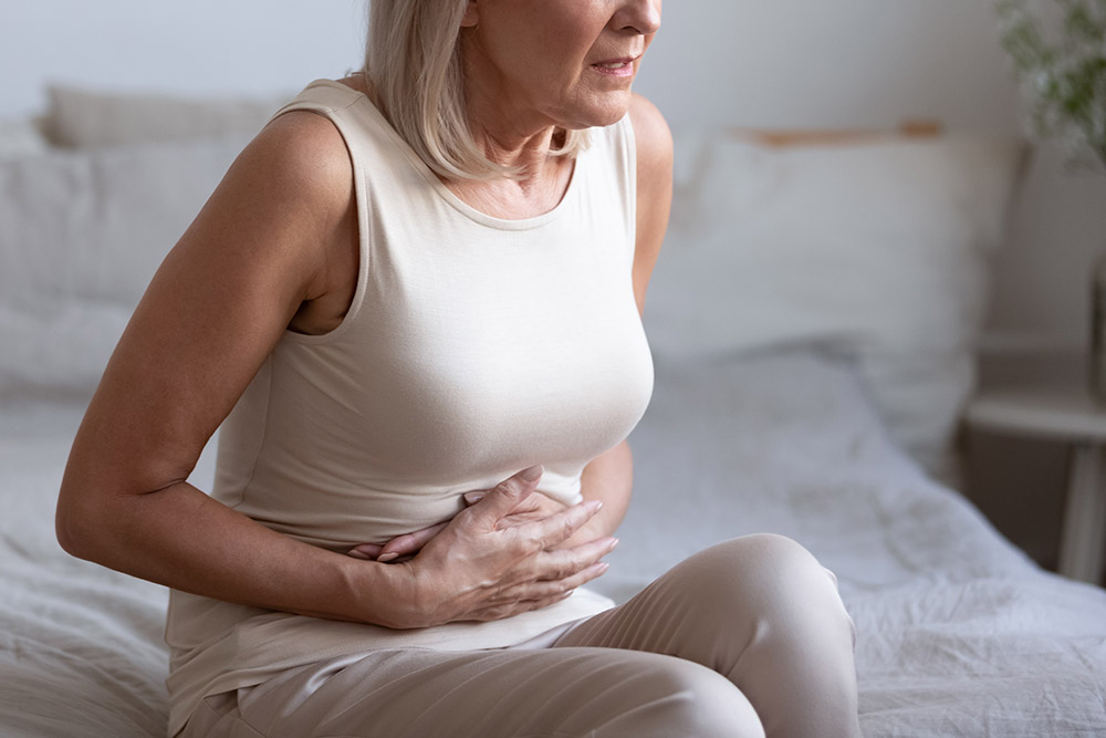 Unhealthy middle aged frowning woman sitting on bed, putting hands on belly, suffering from strong stomach ache. Unhappy older lady having painful feeling in abdomen, pancreatitis gastritis symptom.
