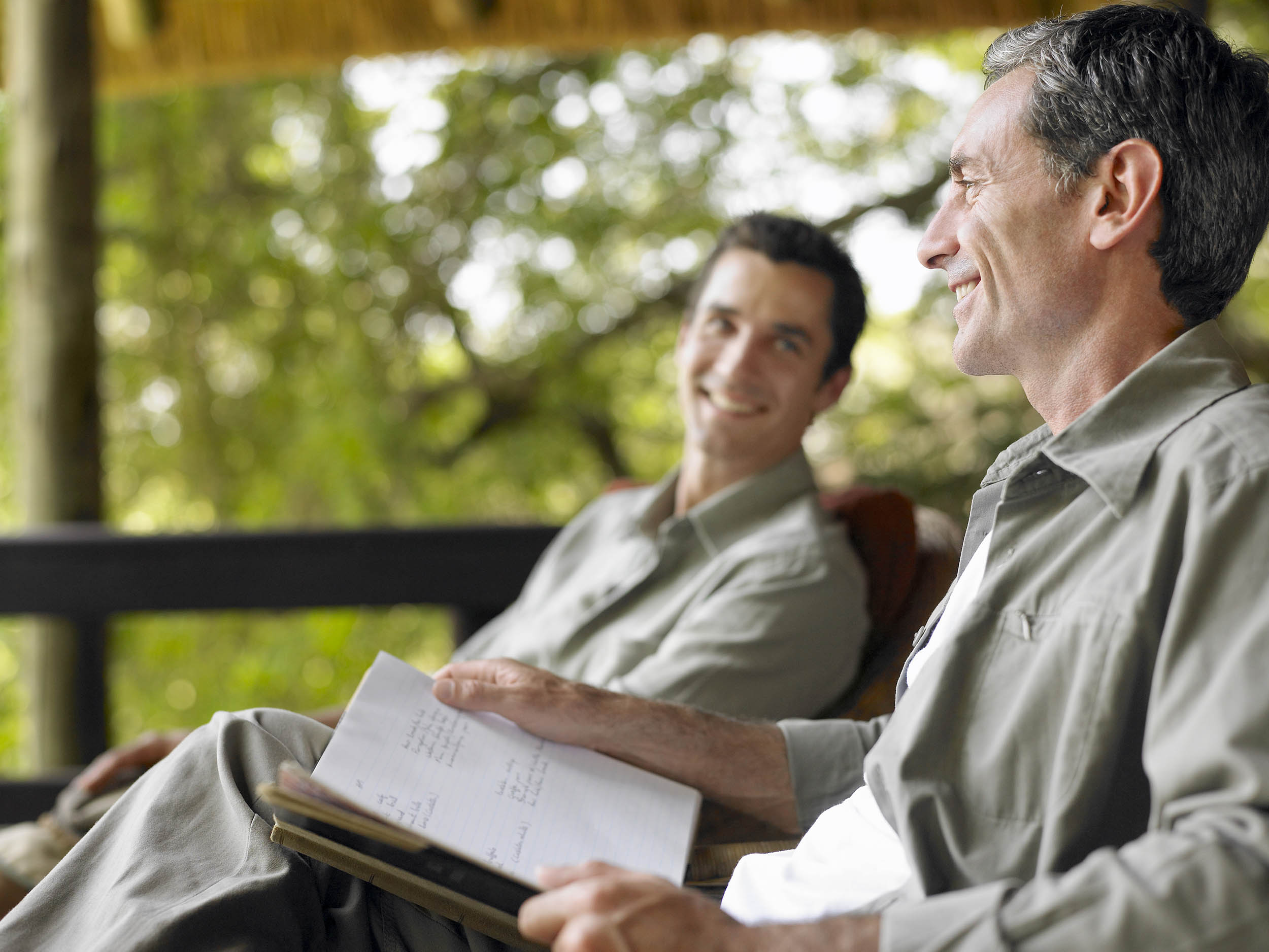 Side view of a happy man with book sitting on terrace with blurred male friend in background