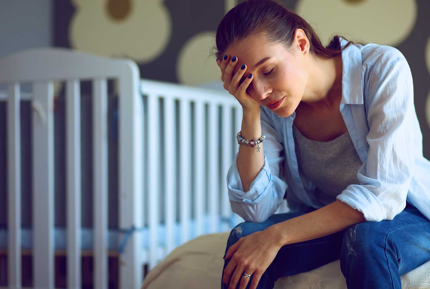 Young tired woman sitting on the bed near children's cot.